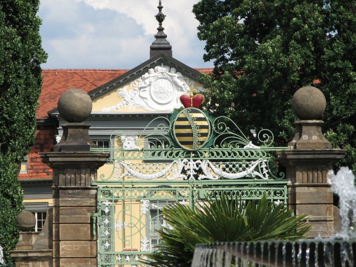 Seit 2003 erstrahlen der Zaun und das mit schmiedeeisernen Akanthusblättern reich verzierte dreiteilige Barockportal der Orangerie wieder in altem Glanz. Gekrönt wird das zweiflügelige Mitteltor seit 1774 vom sächsischen Rautenkranzwappen mit Herzogskrone - hier aufgenommen vor dem Hintergrund des mit dem großen Staatswappen verzierten Giebels des Schlosses Friedrichsthal. (Foto: Cramer)